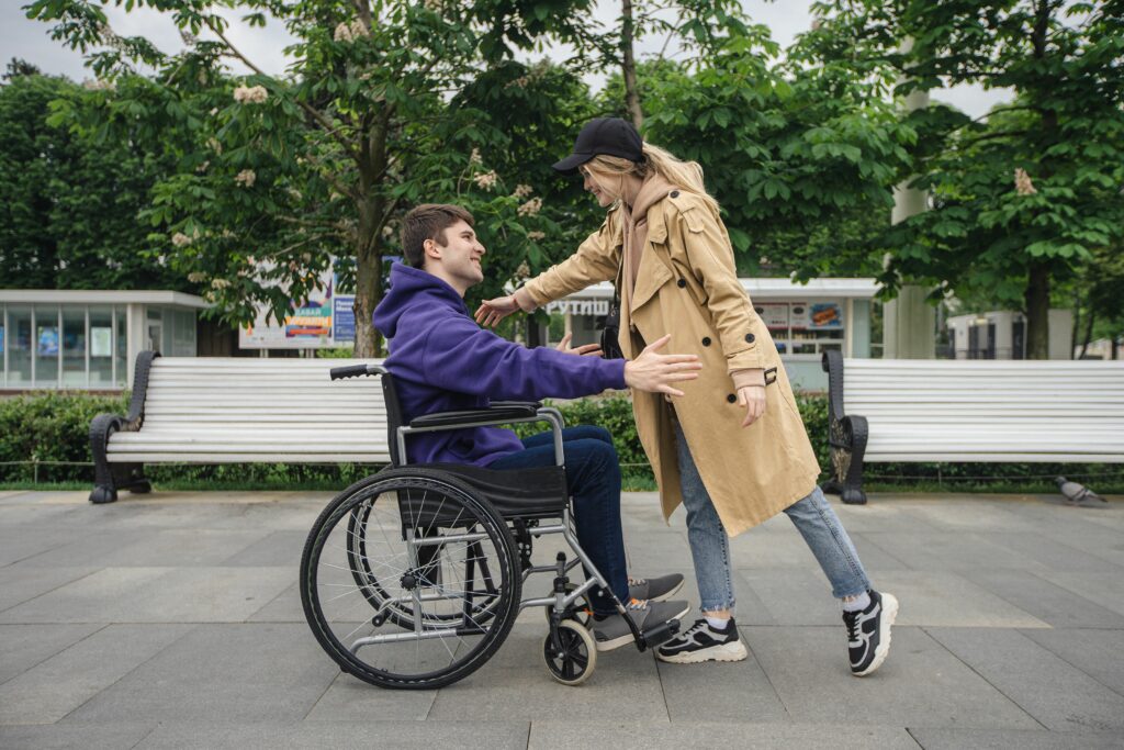 Accueil A man in a wheelchair and a woman greet each other warmly in a park setting.